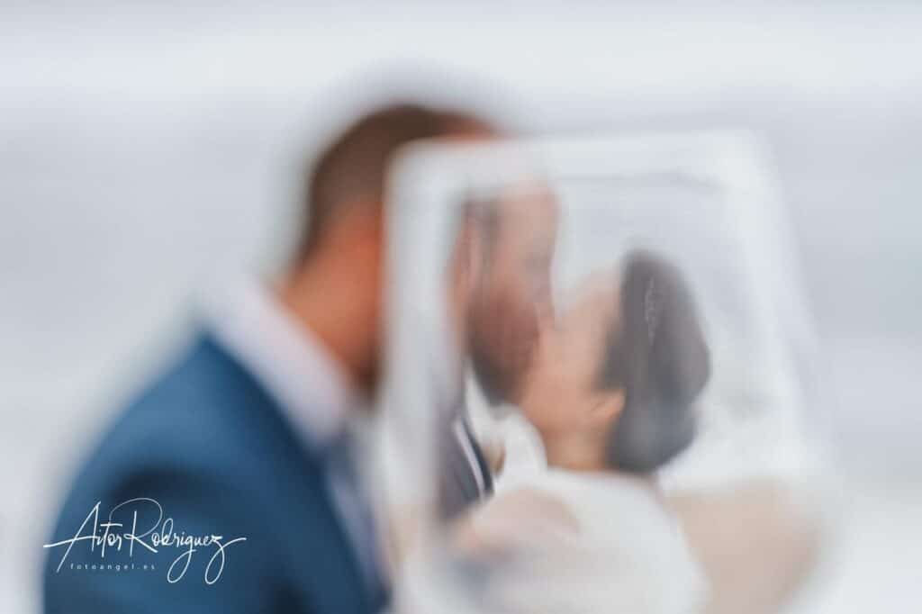 Pareja de novios besándose, capturados a través de una copa de vino, con efecto desenfocado y romántico en su boda en Zarautz.